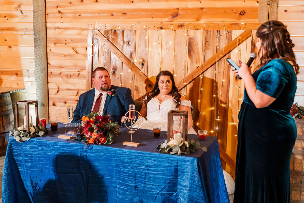 Courtney & John sit at a blue sweetheart table as a bridesmaid gives a toast indoors at Covey Creek.