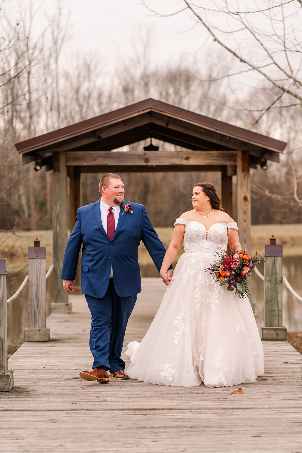Courtney & John smile walking hand in hand on the pond dock past the wooden pavilion at Covey Creek.