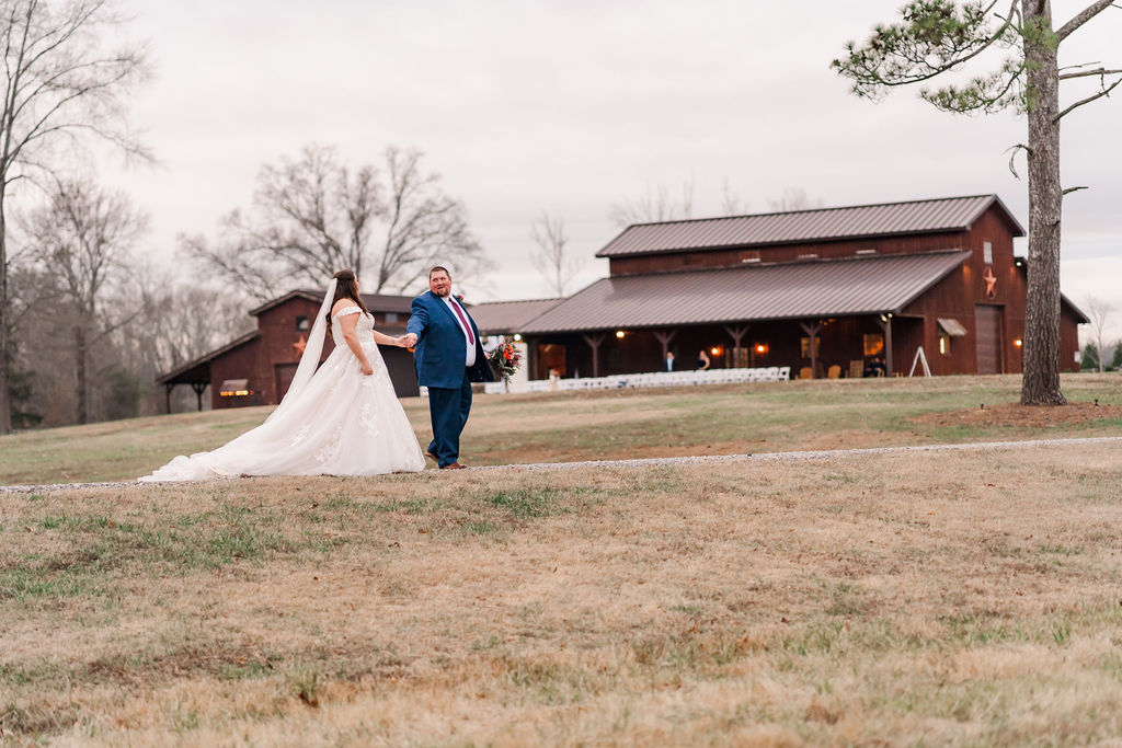 Courtney & John walk a grassy path before the glowing barn porch, her long train trailing at Covey Creek.
