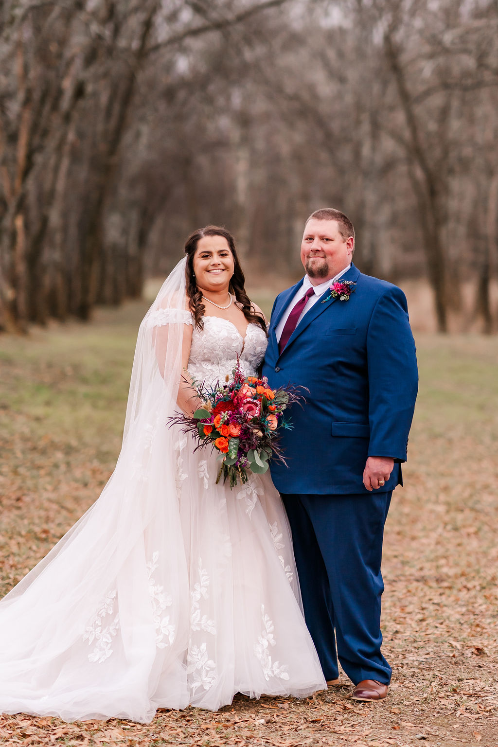 Courtney & John pose among bare winter trees; bride in lace gown with bright bouquet, groom in blue suit at Covey Creek.