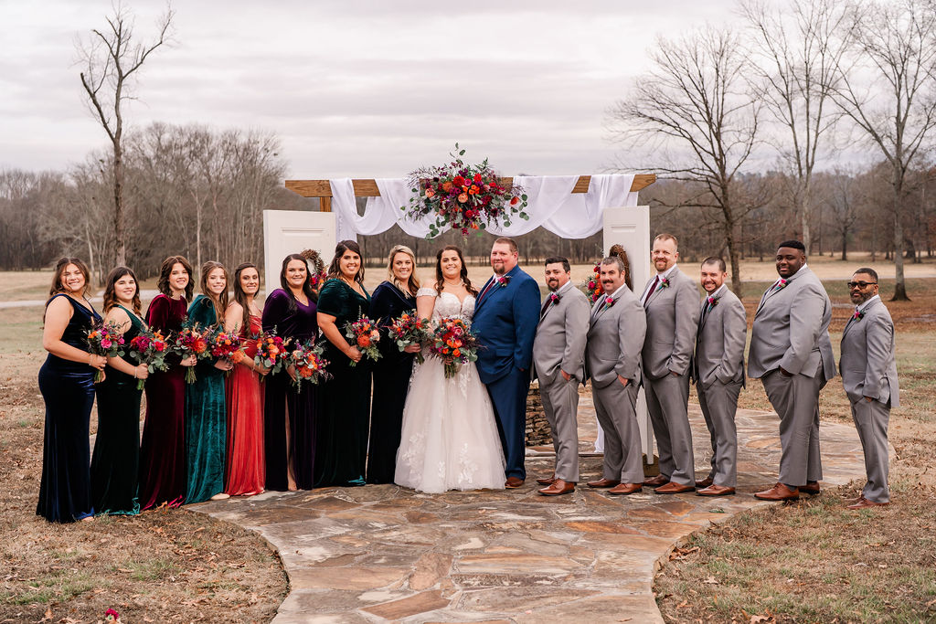 Courtney & John stand with their wedding party under a flower-draped arbor with white doors at Covey Creek.