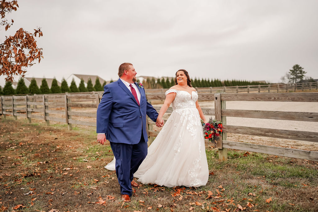 Courtney & John walk hand in hand by a wooden fence, bride in lace gown with a bright bouquet at Covey Creek.