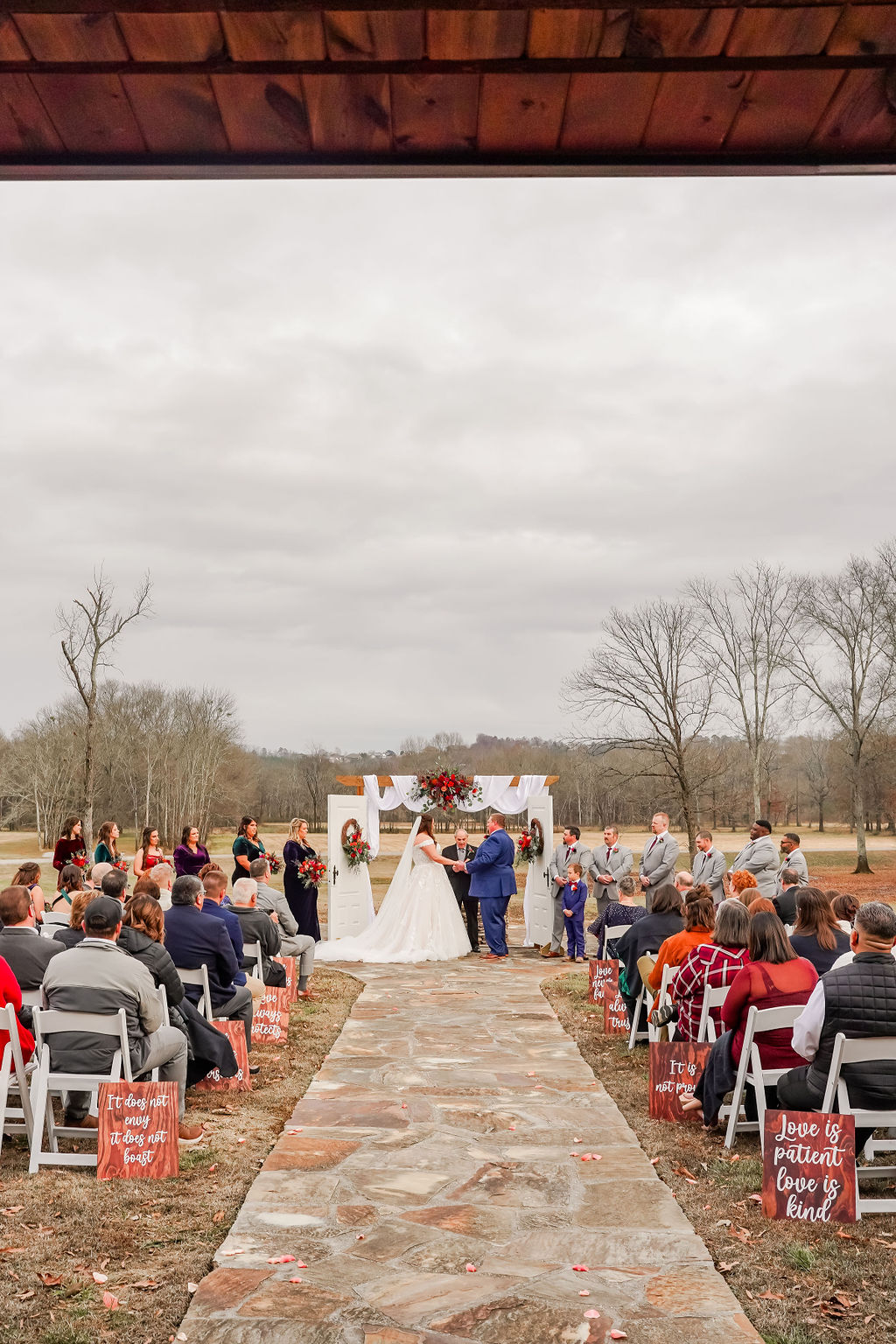 View down the stone aisle as Courtney & John stand at the flower-draped arbor with guests at Covey Creek.
