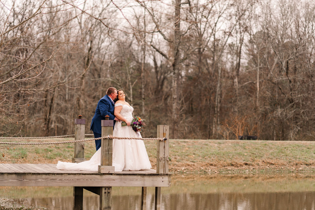 Courtney & John pose on the wooden pond dock; bride in lace gown with an orange bouquet at Covey Creek.