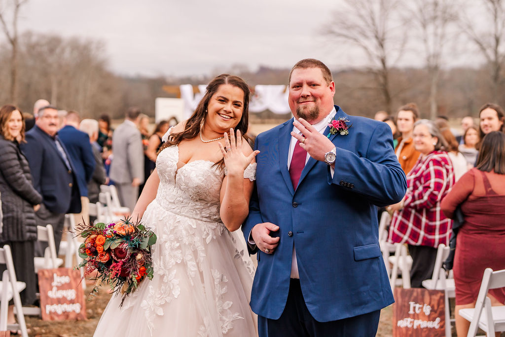 Courtney & John flash their new wedding rings, smiling down the outdoor ceremony aisle at Covey Creek.