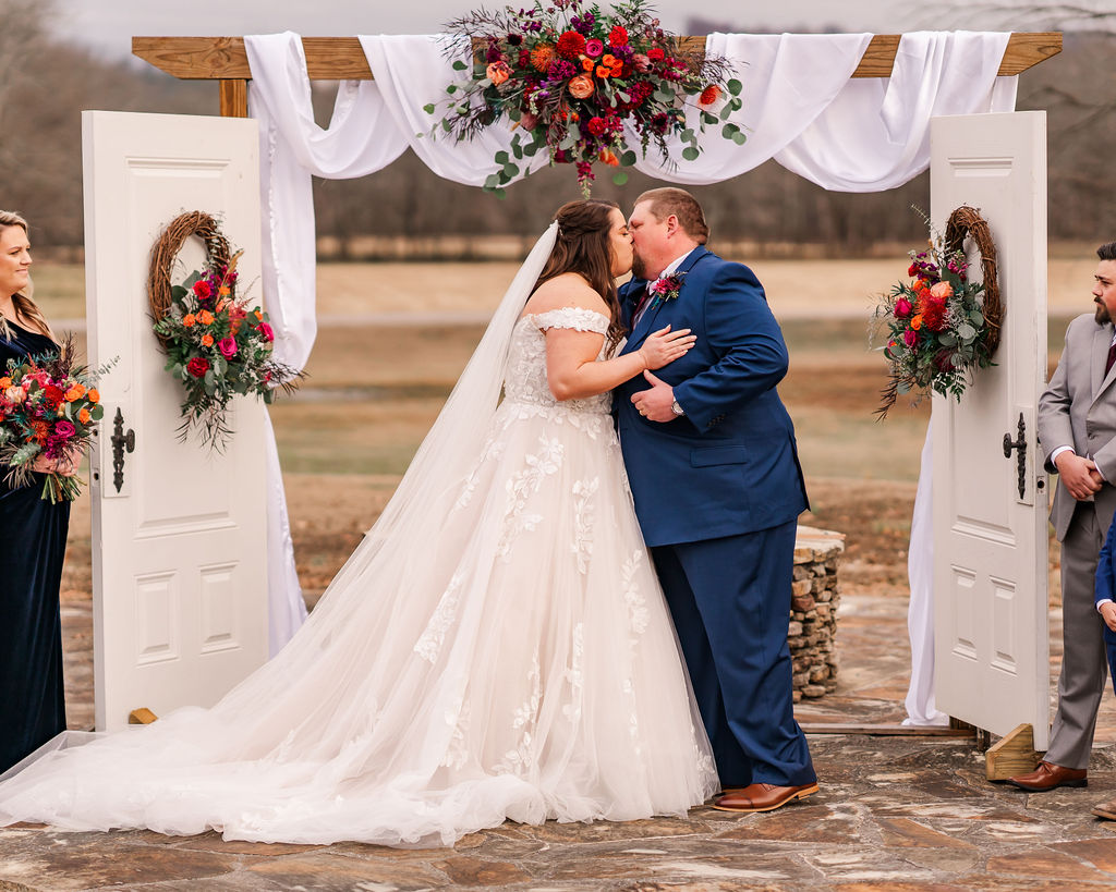 Courtney & John share a first kiss under a flower-draped arbor with white doors on the patio at Covey Creek.