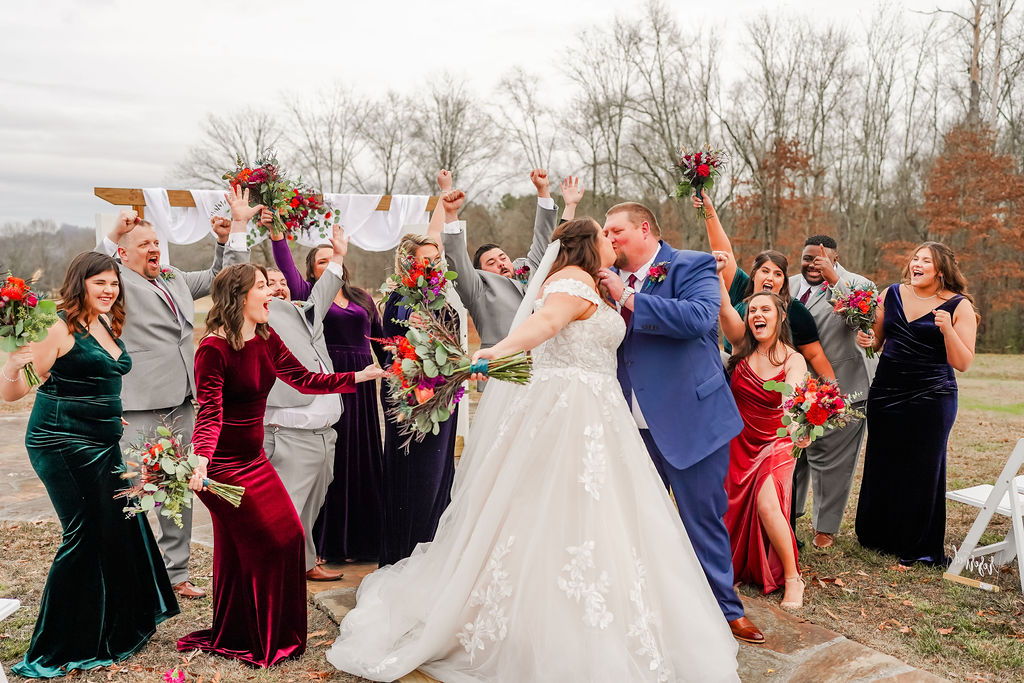 Courtney & John kiss as their jewel-toned wedding party cheers around them outdoors at Covey Creek.