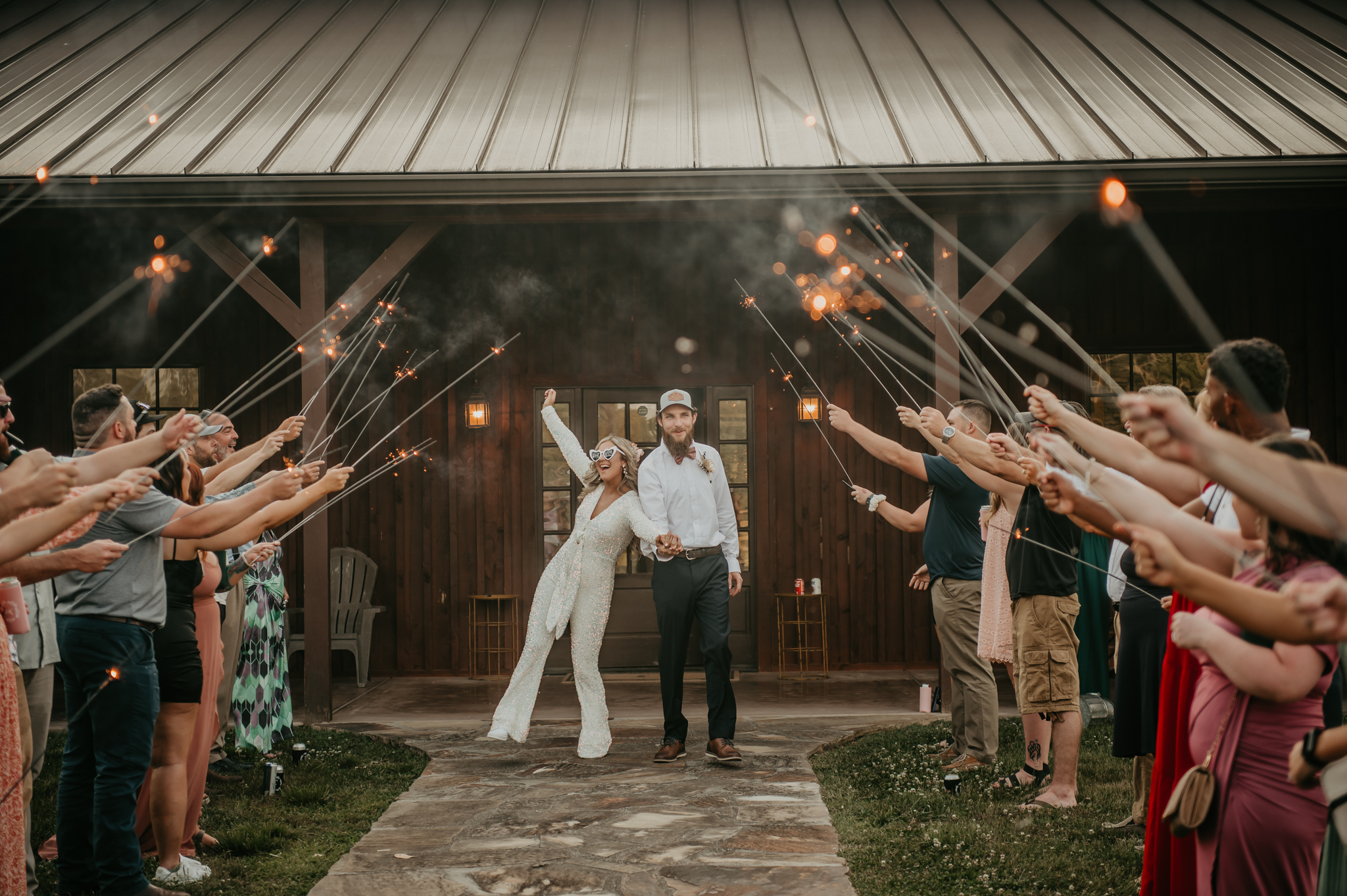Hali & Kyle exit through sparklers at Covey Creek; bride in sequined jumpsuit and heart sunglasses.