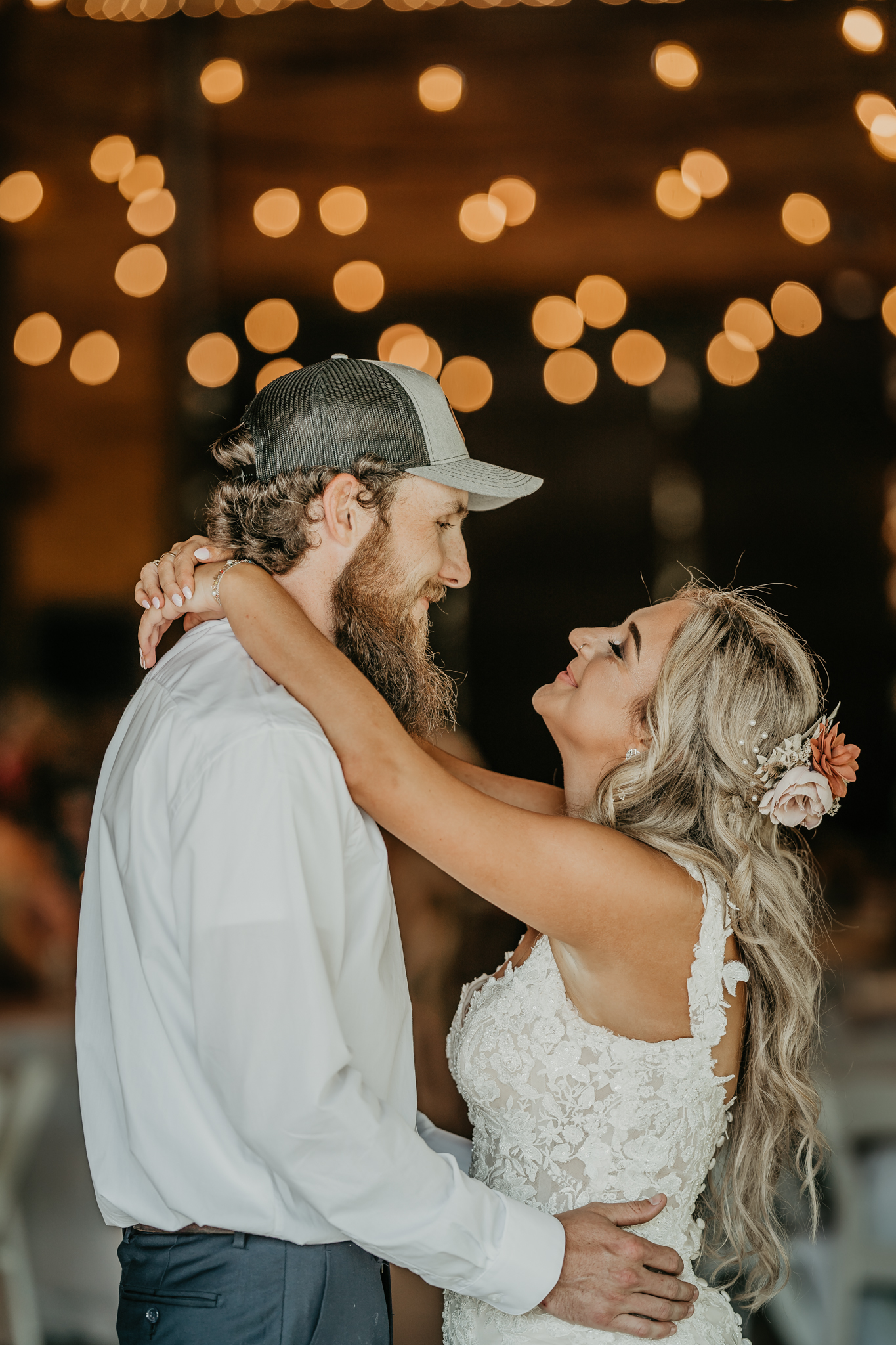 Hali & Kyle embrace under warm string lights indoors at Covey Creek, bride in lace with floral hairpins.