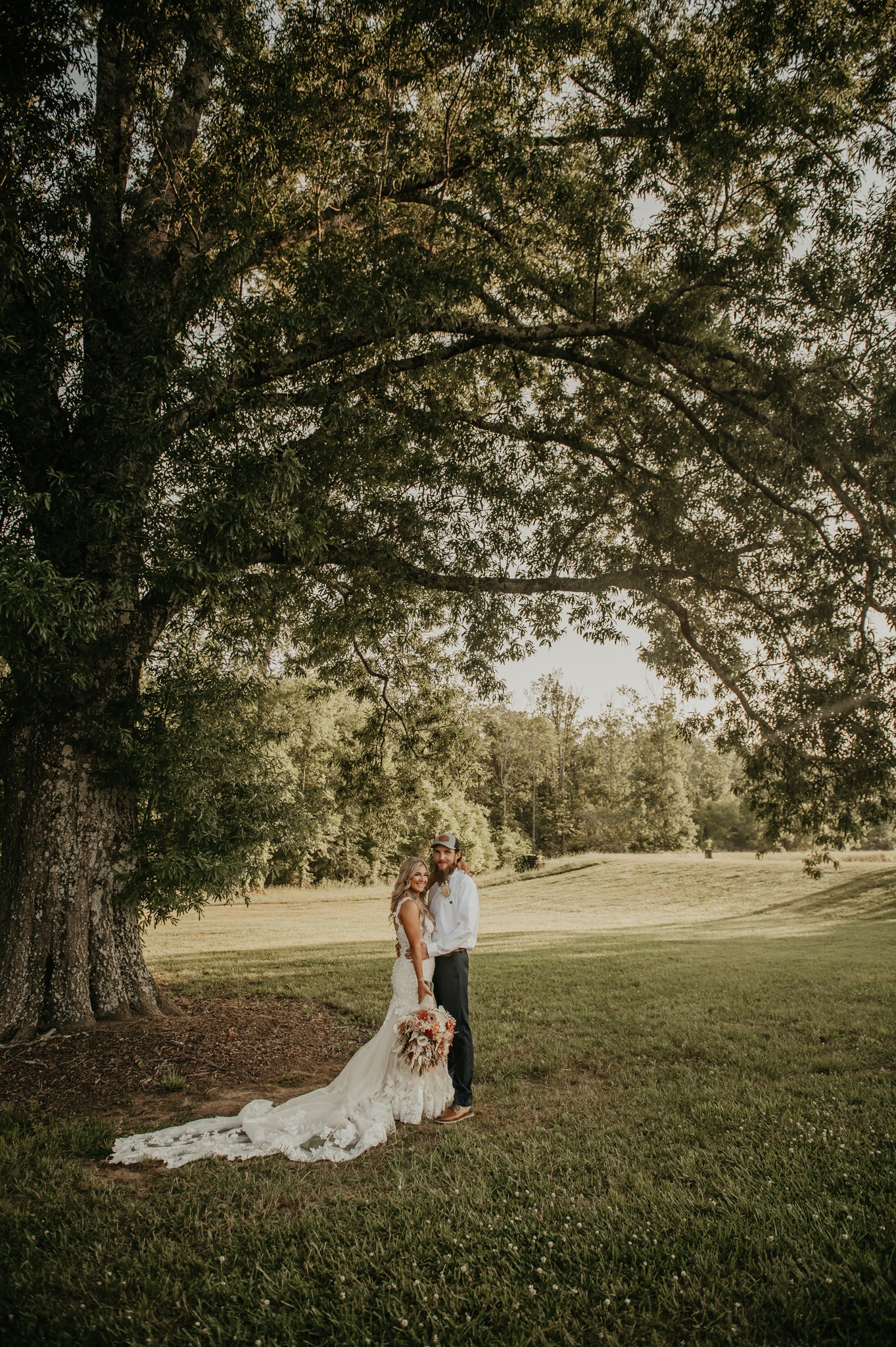 Hali & Kyle stand under a large tree in a grassy field at Covey Creek, her lace train spread on the grass.