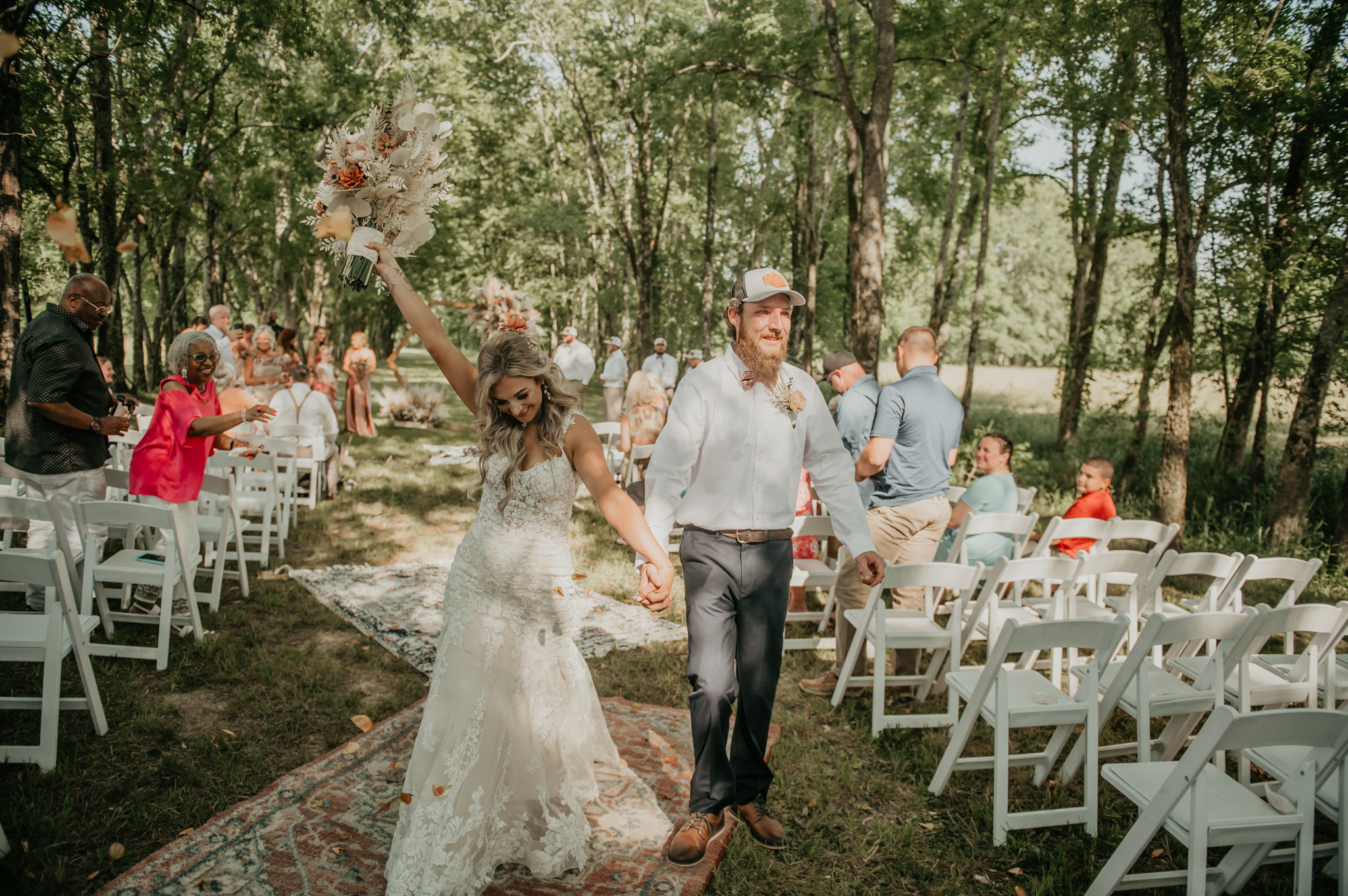 Hali & Kyle walk the rug aisle at Covey Creek as she raises her bouquet, guests cheering under the trees.