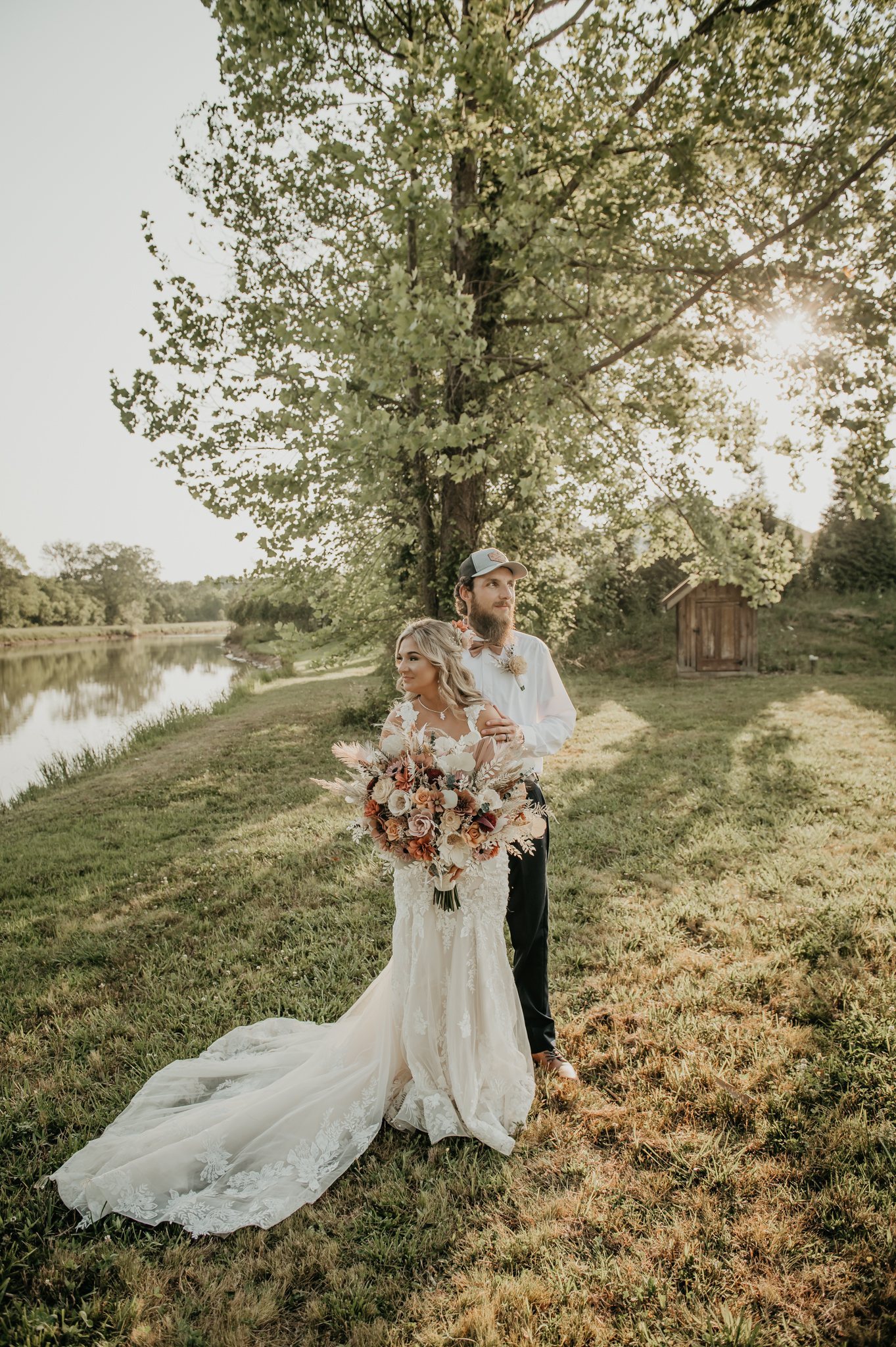 Hali & Kyle pose by the pond at Covey Creek, lace train on the grass and warm florals in her bouquet.