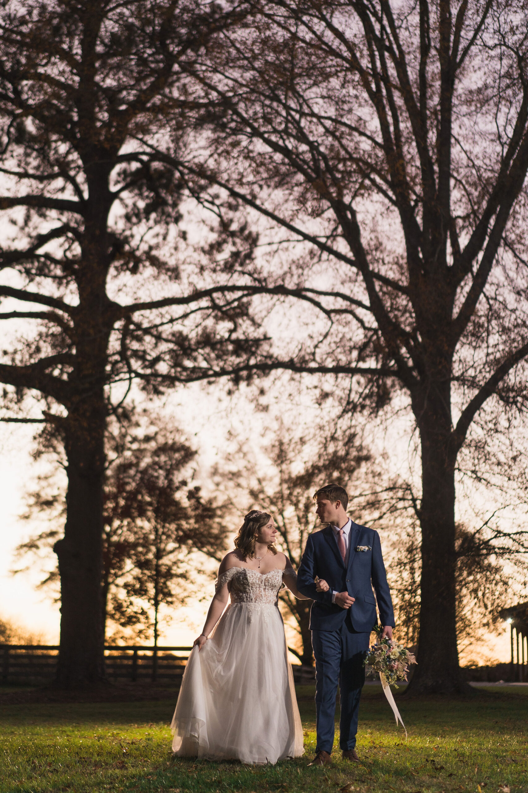 Ana & Carter walk arm in arm at twilight between bare trees on the grassy field at Covey Creek