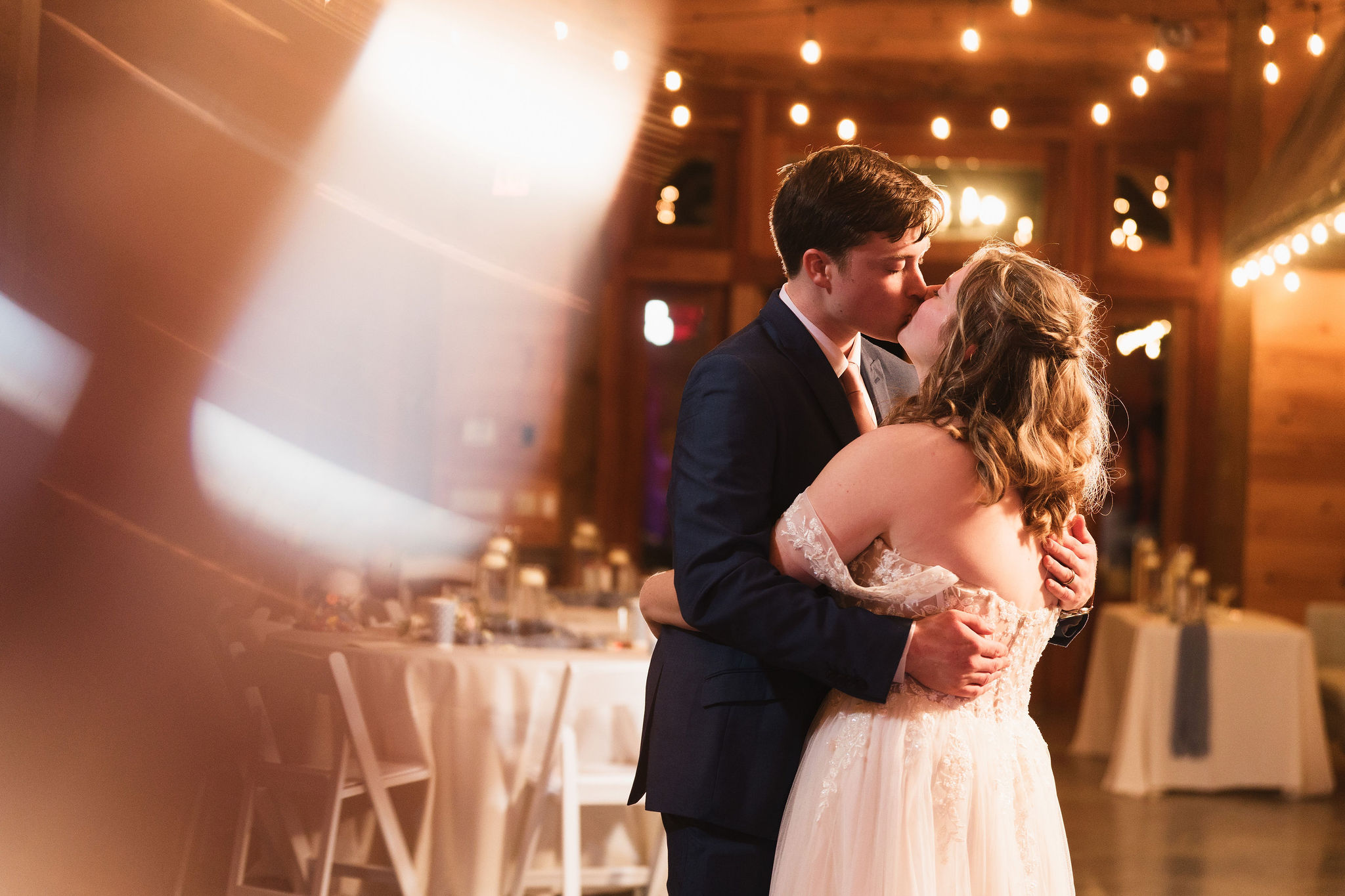 Ana & Carter kiss during their reception dance amid warm string-light bokeh in the barn at Covey Creek
