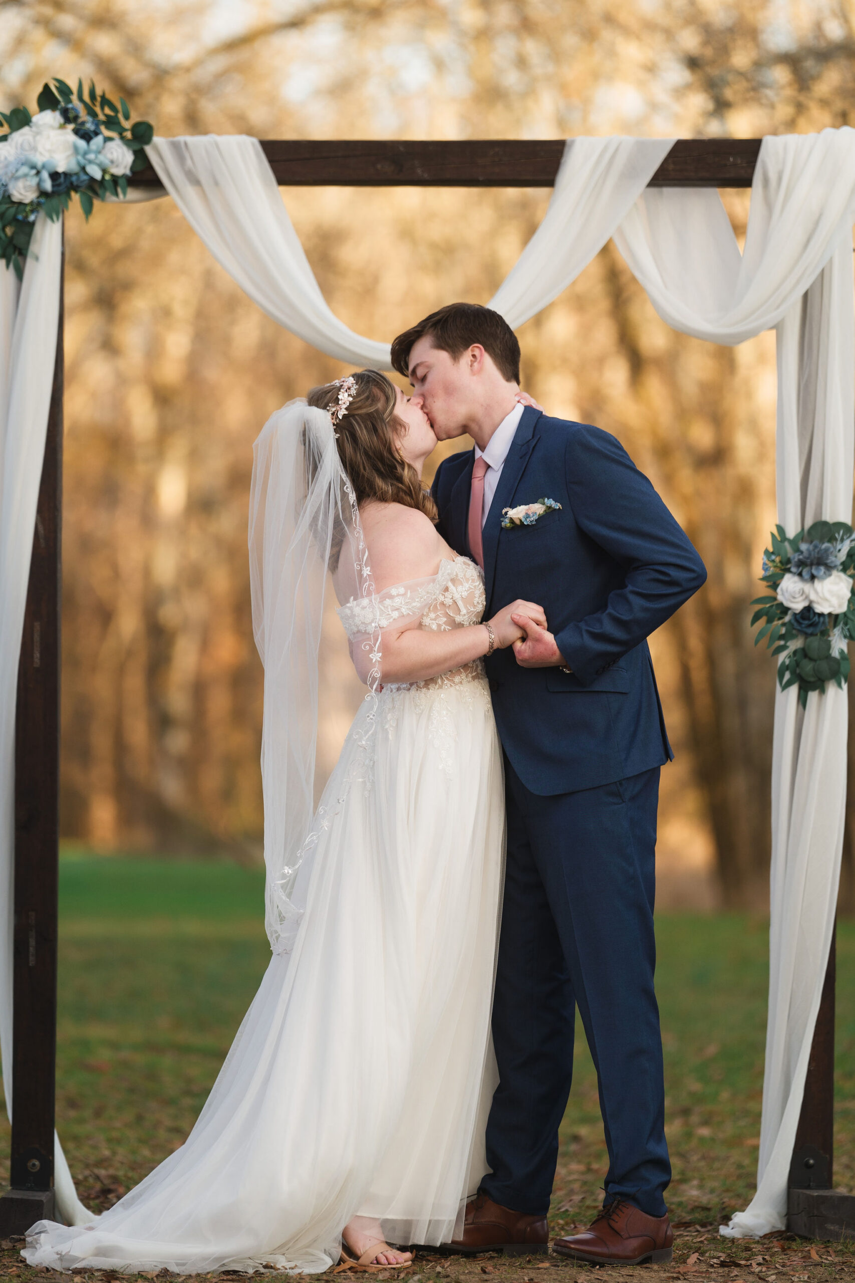 Ana & Carter kiss under a white-draped wooden arch with blue florals outdoors at Covey Creek