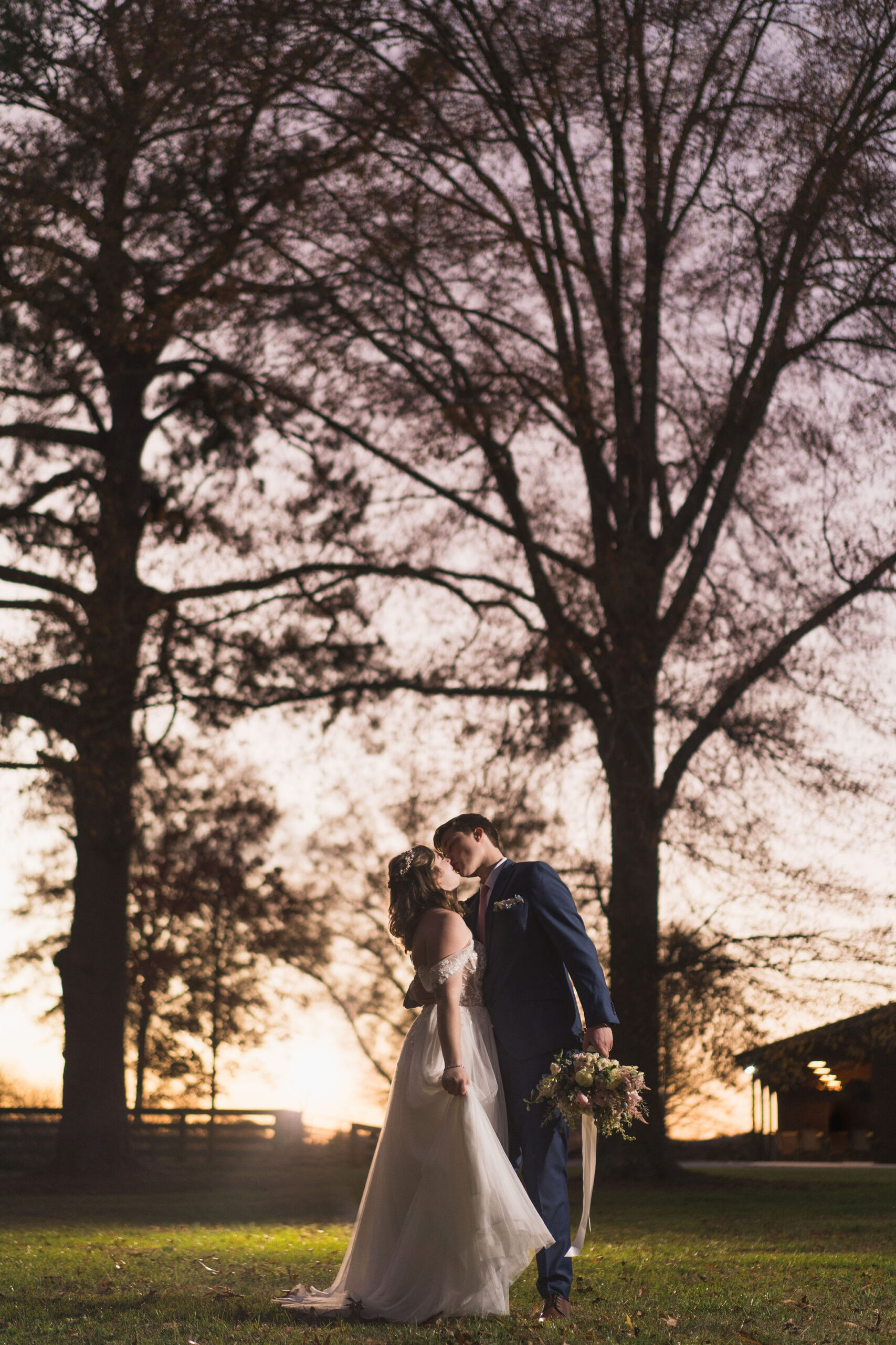 Ana & Carter kiss at dusk between towering trees, bouquet in hand, on the grassy field at Covey Creek