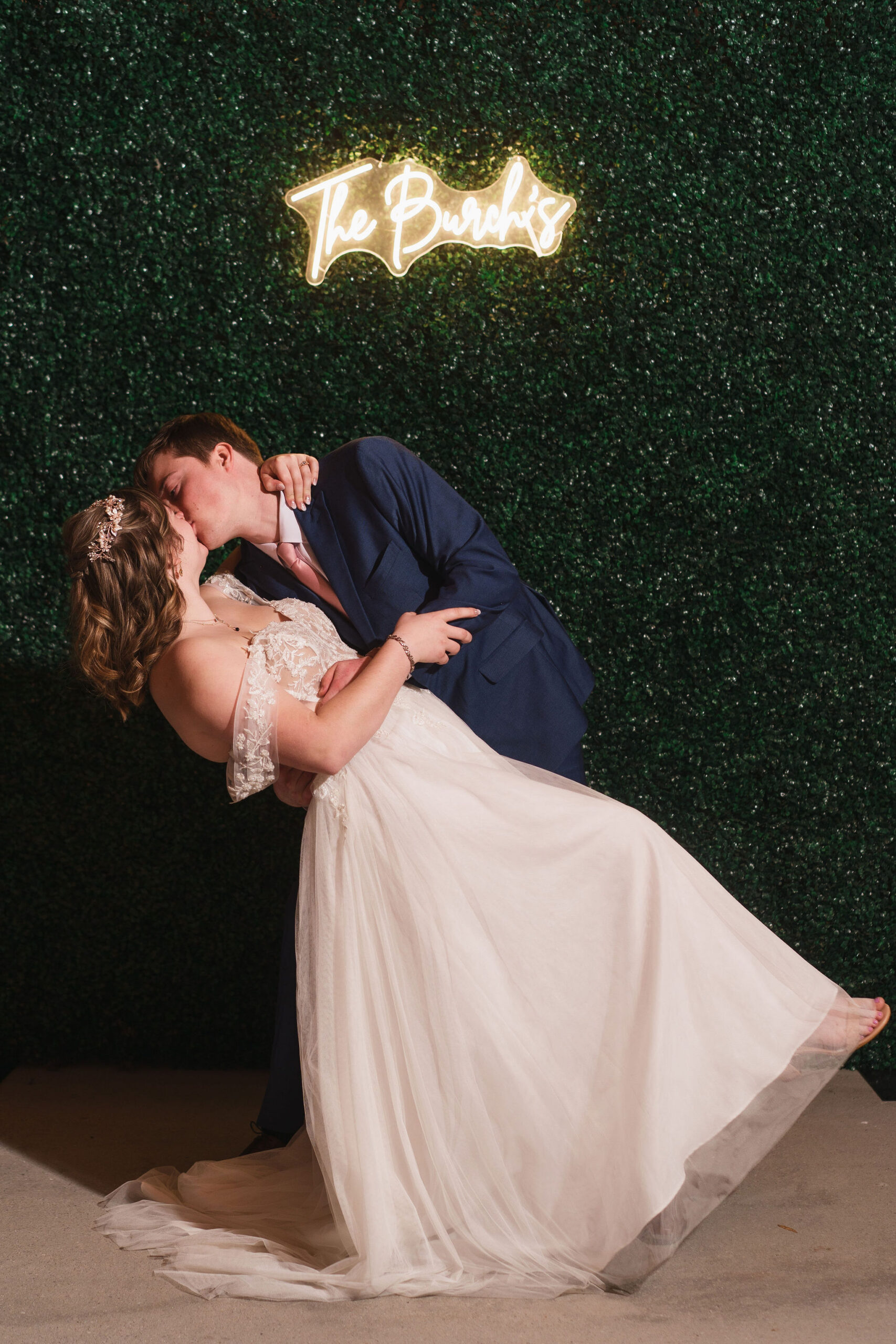 Ana & Carter dip and kiss before a greenery wall and The Burch's neon sign indoors at Covey Creek