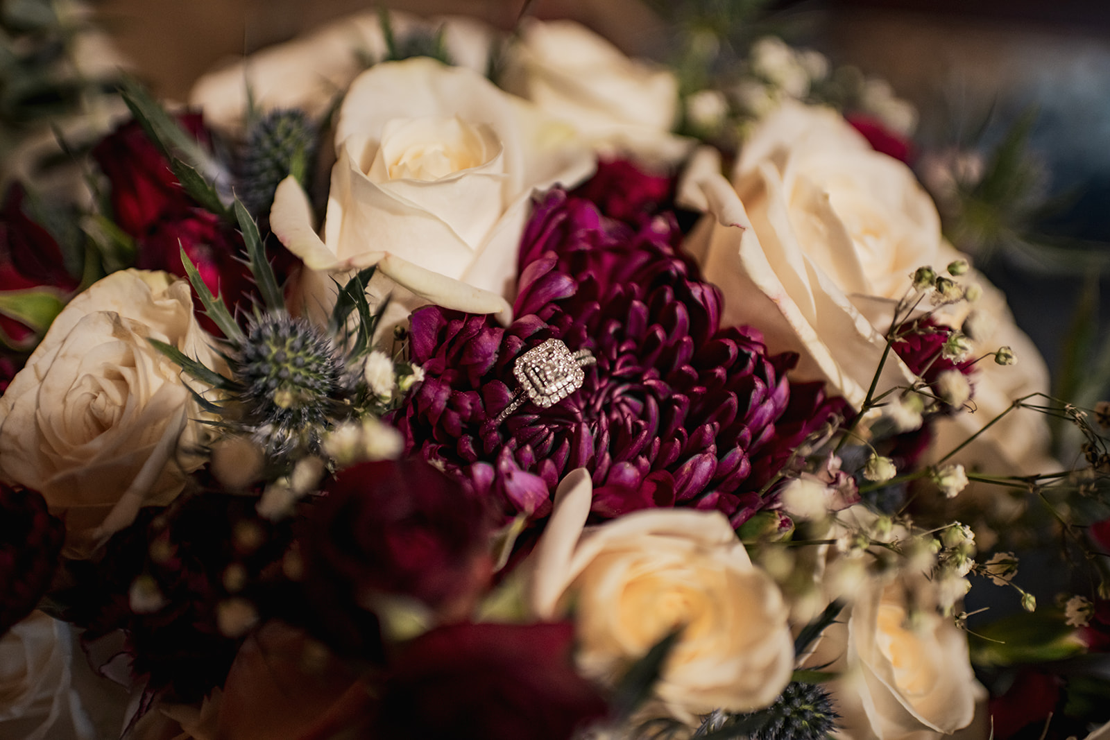 Halo diamond ring rests on a plum chrysanthemum amid ivory roses and blue thistle from Annie & Will’s Covey Creek wedding.