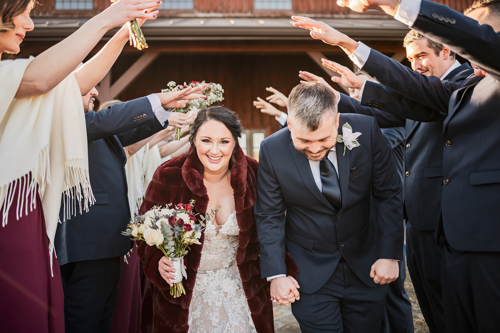 Annie & Will walk a wedding-party arch at Covey Creek, Annie smiling in a burgundy fur coat with white rose bouquet.