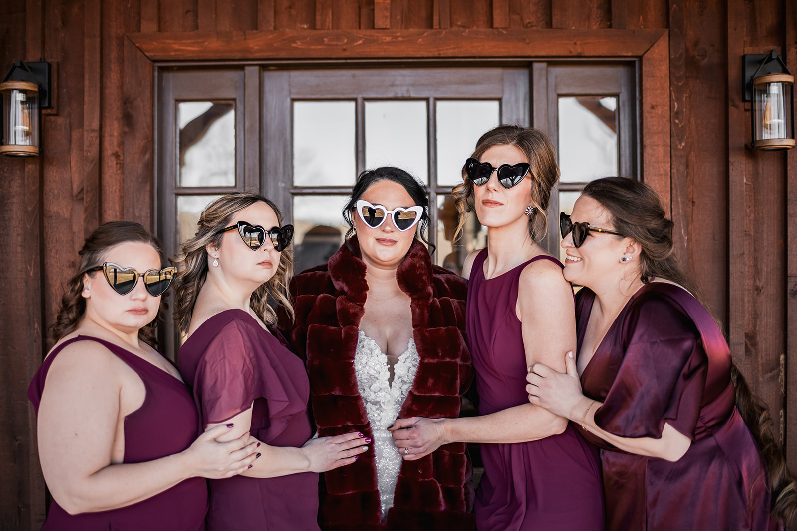 Annie & Will’s bridal party: Annie and burgundy bridesmaids in heart sunglasses before Covey Creek’s wood siding and lanterns.