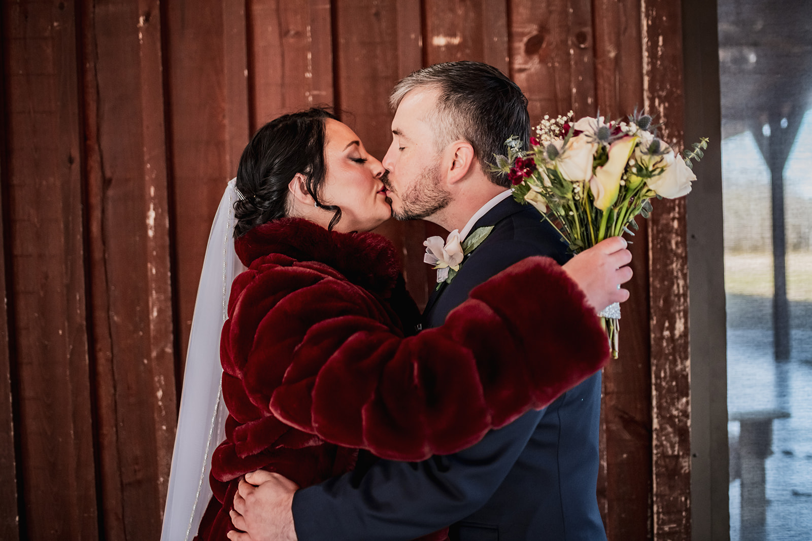 Annie & Will kiss against Covey Creek’s red barn wall, Annie in a burgundy fur wrap holding white roses and blue thistle.