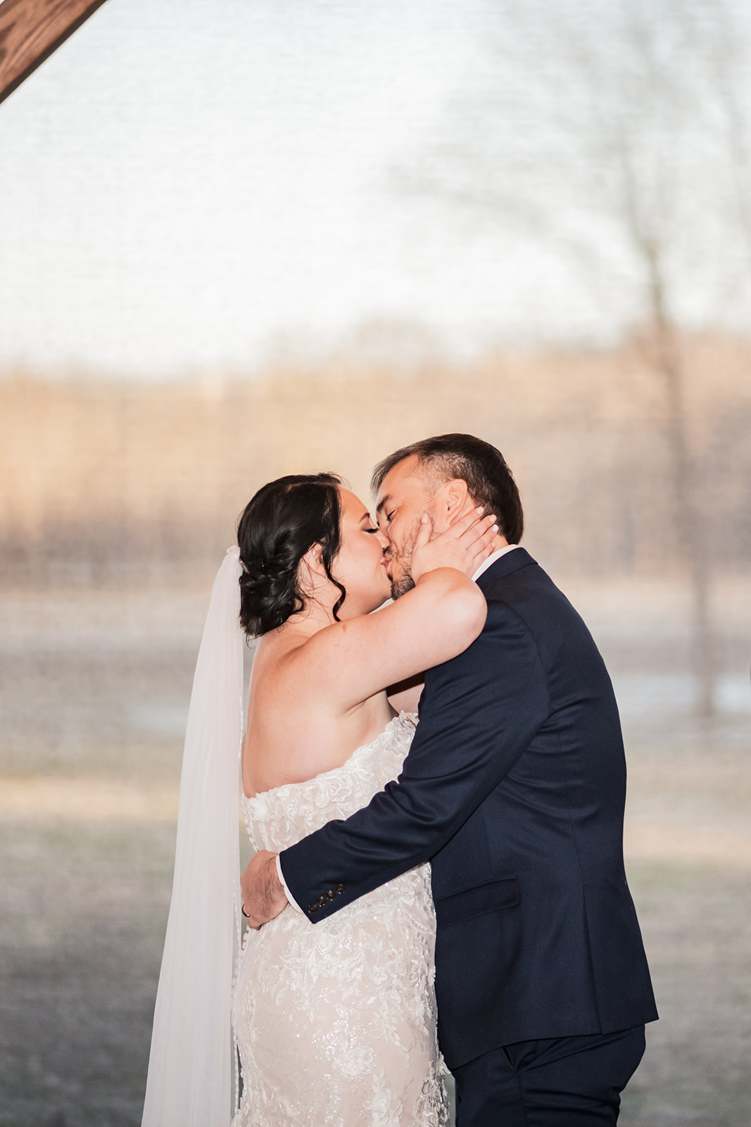 Annie & Will kiss under a timber beam at Covey Creek, her lace gown and veil lit against bare winter trees beyond.