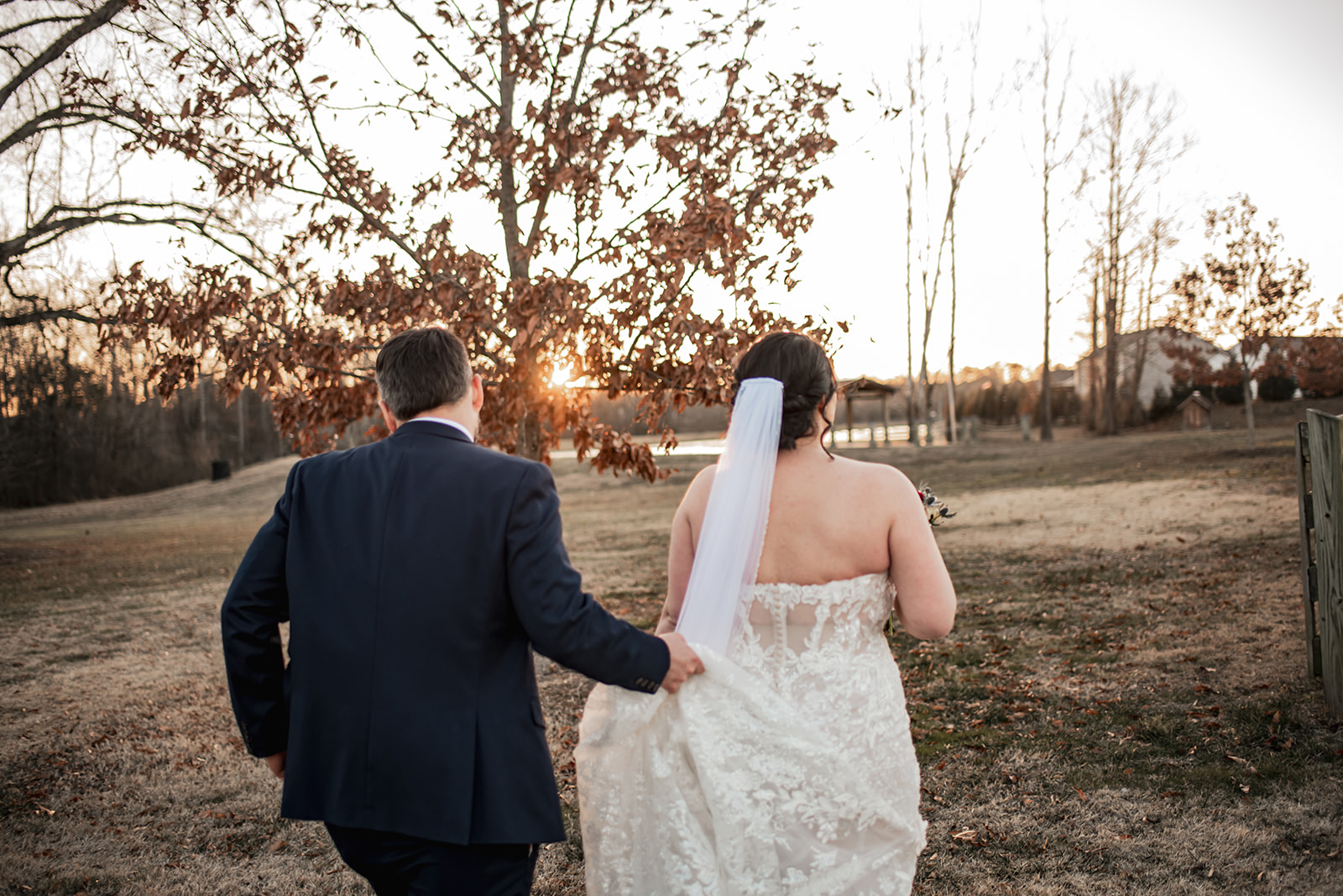 Annie & Will walk toward Covey Creek’s pond and gazebo at golden hour as Will carries her lace train through the grass.