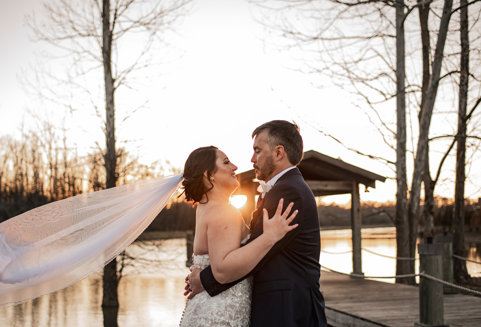 Annie & Will embrace on Covey Creek’s pond dock at sunset, her veil blowing as bare trees stand in the golden water.