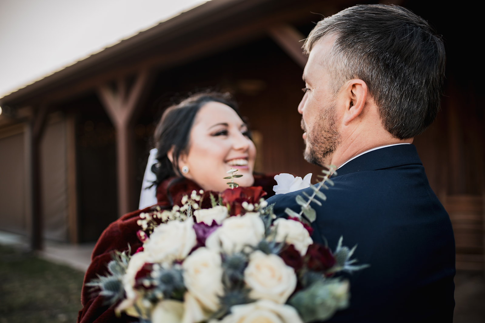 Annie smiles over Will’s shoulder in a burgundy wrap at Covey Creek for Annie & Will, rose bouquet soft in the foreground.