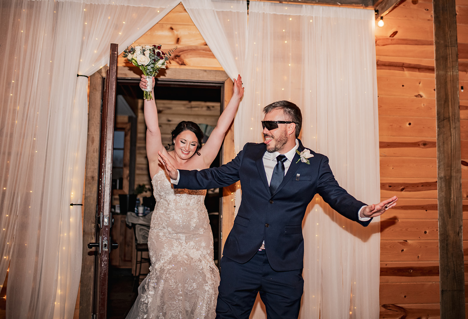 Annie & Will burst through lit white curtains at Covey Creek, bouquet raised and Will in sunglasses mid-celebration.
