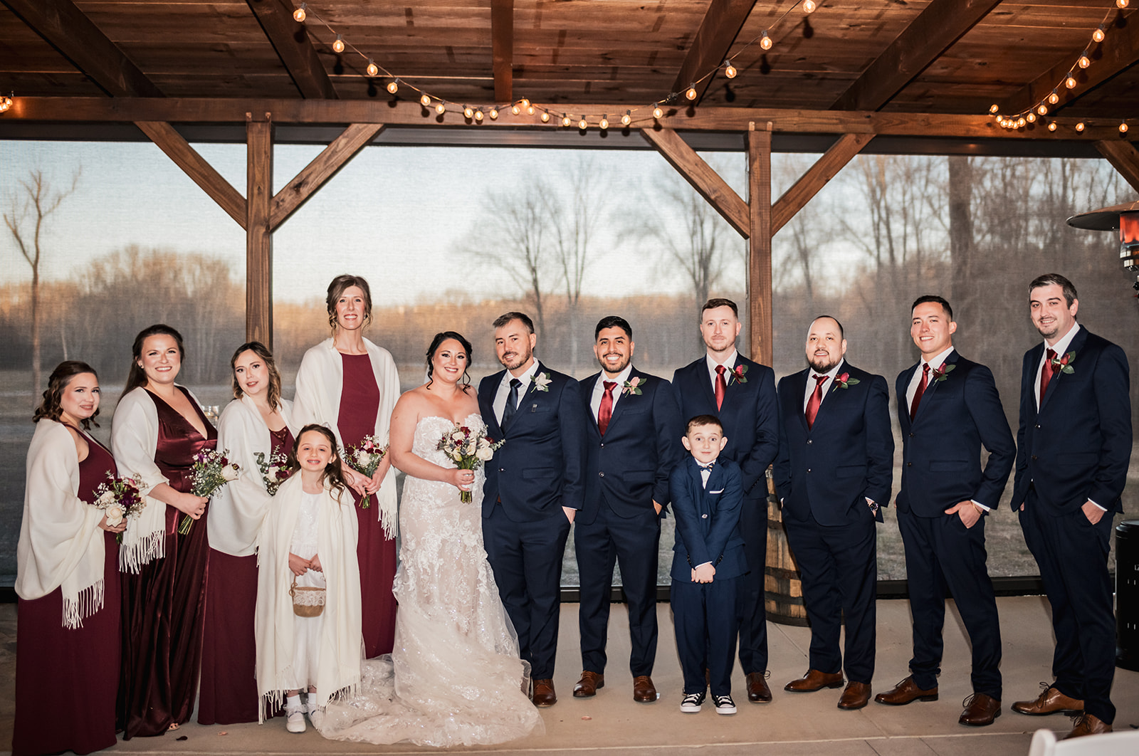 Annie & Will stand centered with burgundy bridesmaids and navy groomsmen under Covey Creek string lights at dusk.