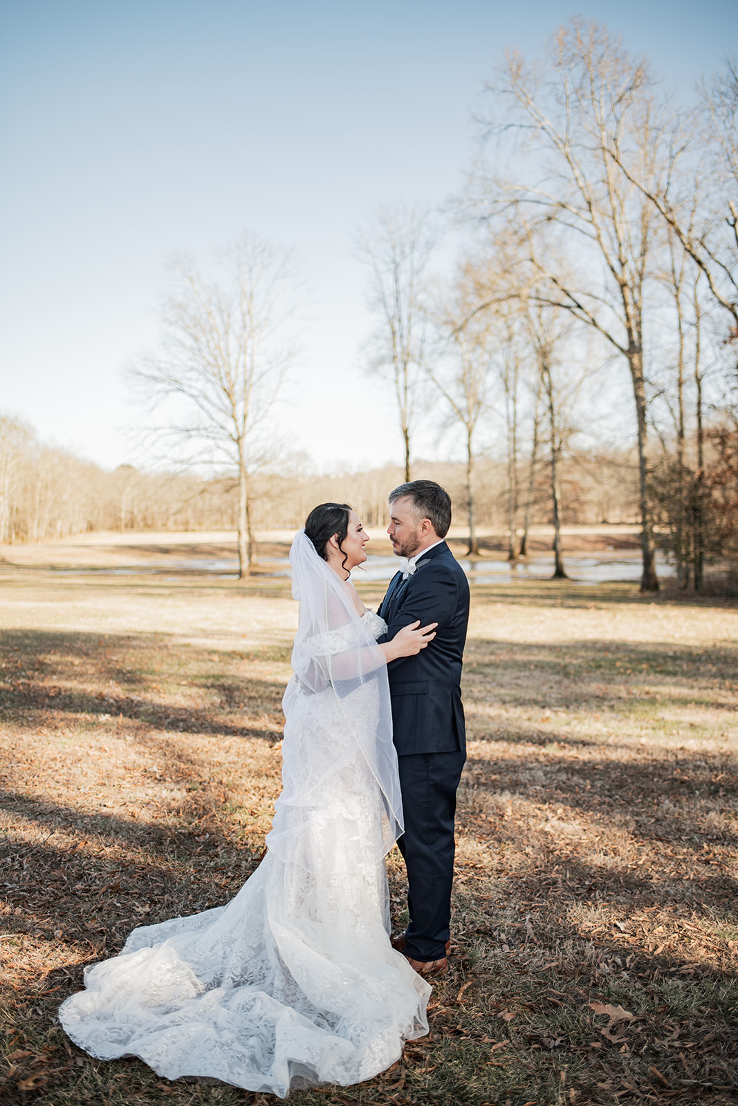 Annie & Will embrace in profile in a winter field at Covey Creek, her lace train spread on dormant grass near the pond.