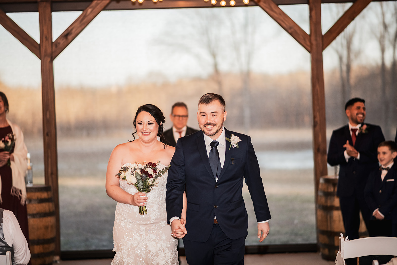 Annie & Will recess hand in hand down Covey Creek’s timber aisle, smiling with white-and-burgundy roses as guests clap.
