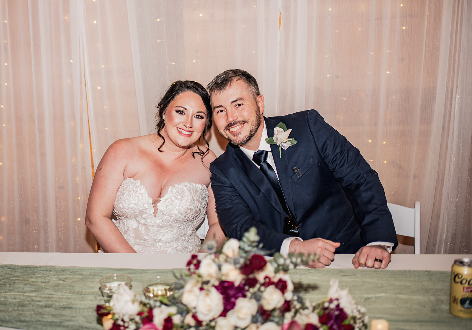 Annie & Will smile at their sweetheart table at Covey Creek, fairy-lit curtain and white-burgundy florals in front.