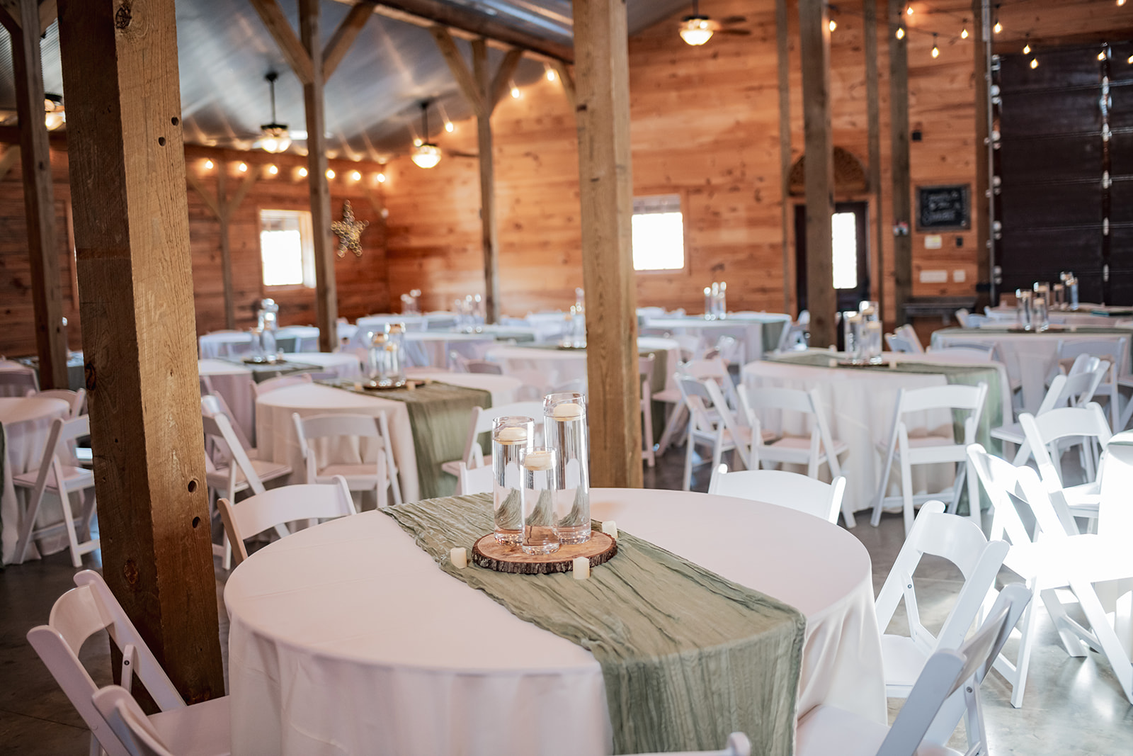 Sage runners and floating candle centerpieces dress round tables under string lights in Covey Creek’s barn for Annie & Will.