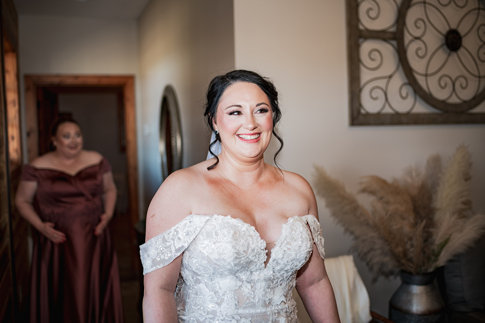 Annie smiles in her off-shoulder lace gown getting ready at Covey Creek for Annie & Will’s day, burgundy dress soft behind.
