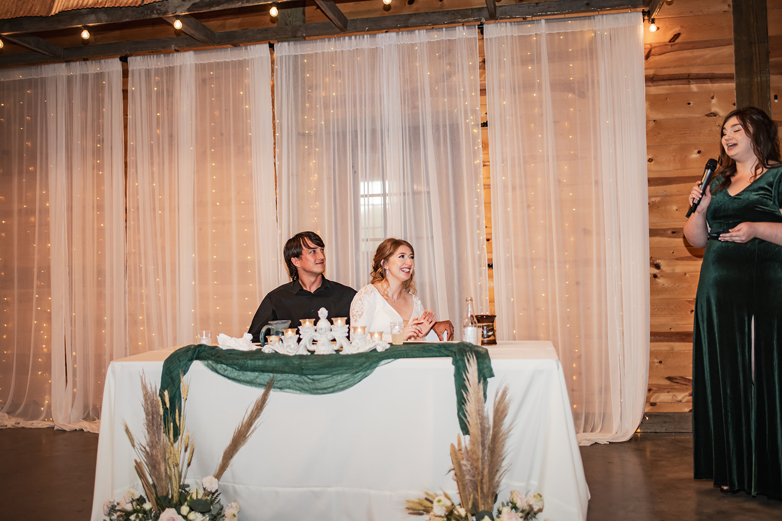 Isabelle & Alex sit at their sweetheart table as a bridesmaid toasts in the Covey Creek barn reception.