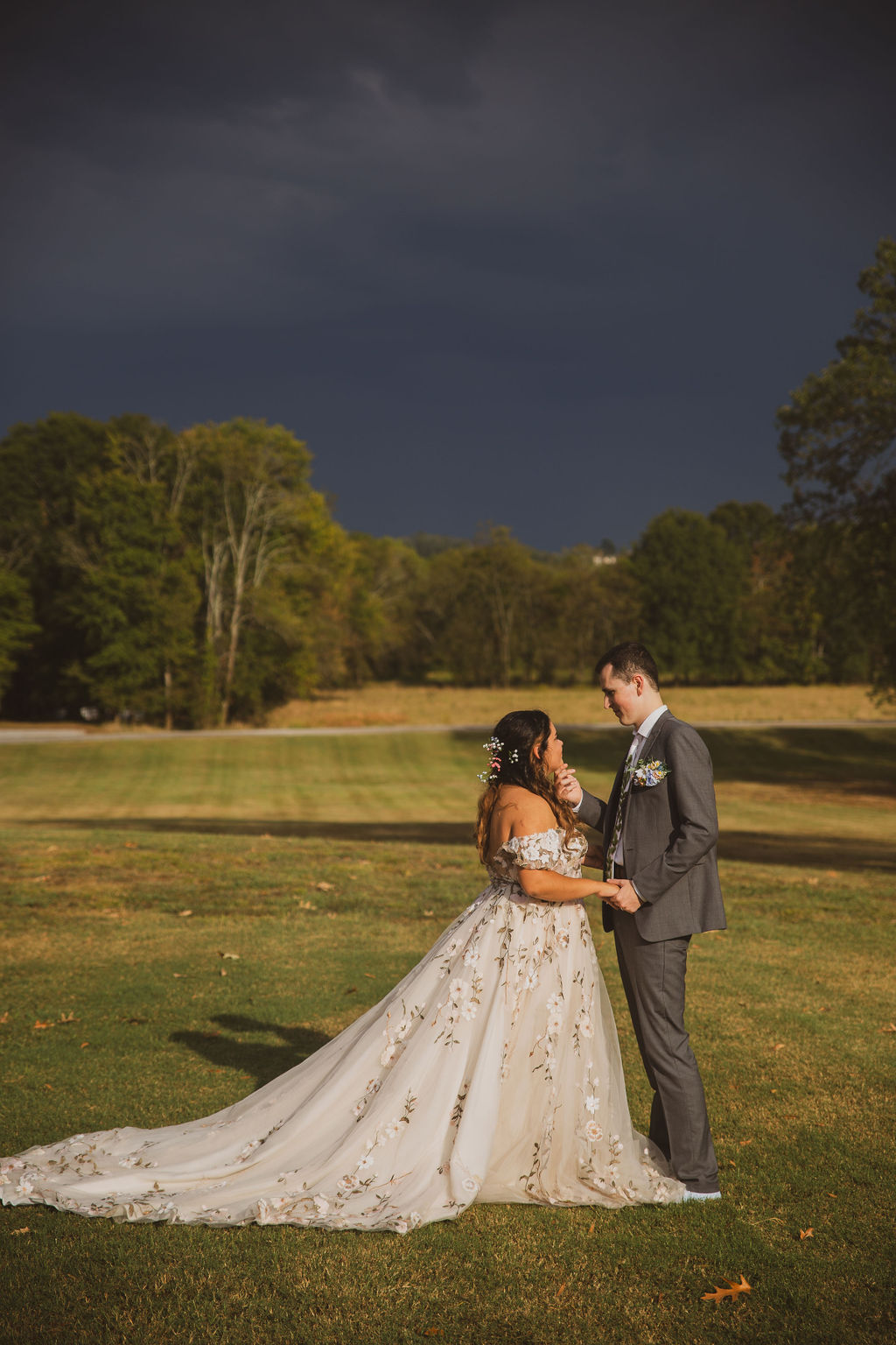Jen & Alec hold each other in a grassy field under a stormy sky in floral gown and grey suit at Covey Creek