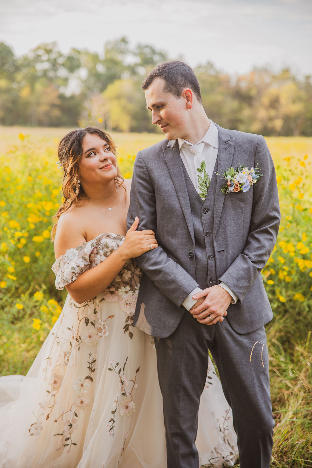 Jen & Alec smile close together among yellow wildflowers in floral gown and grey suit at Covey Creek