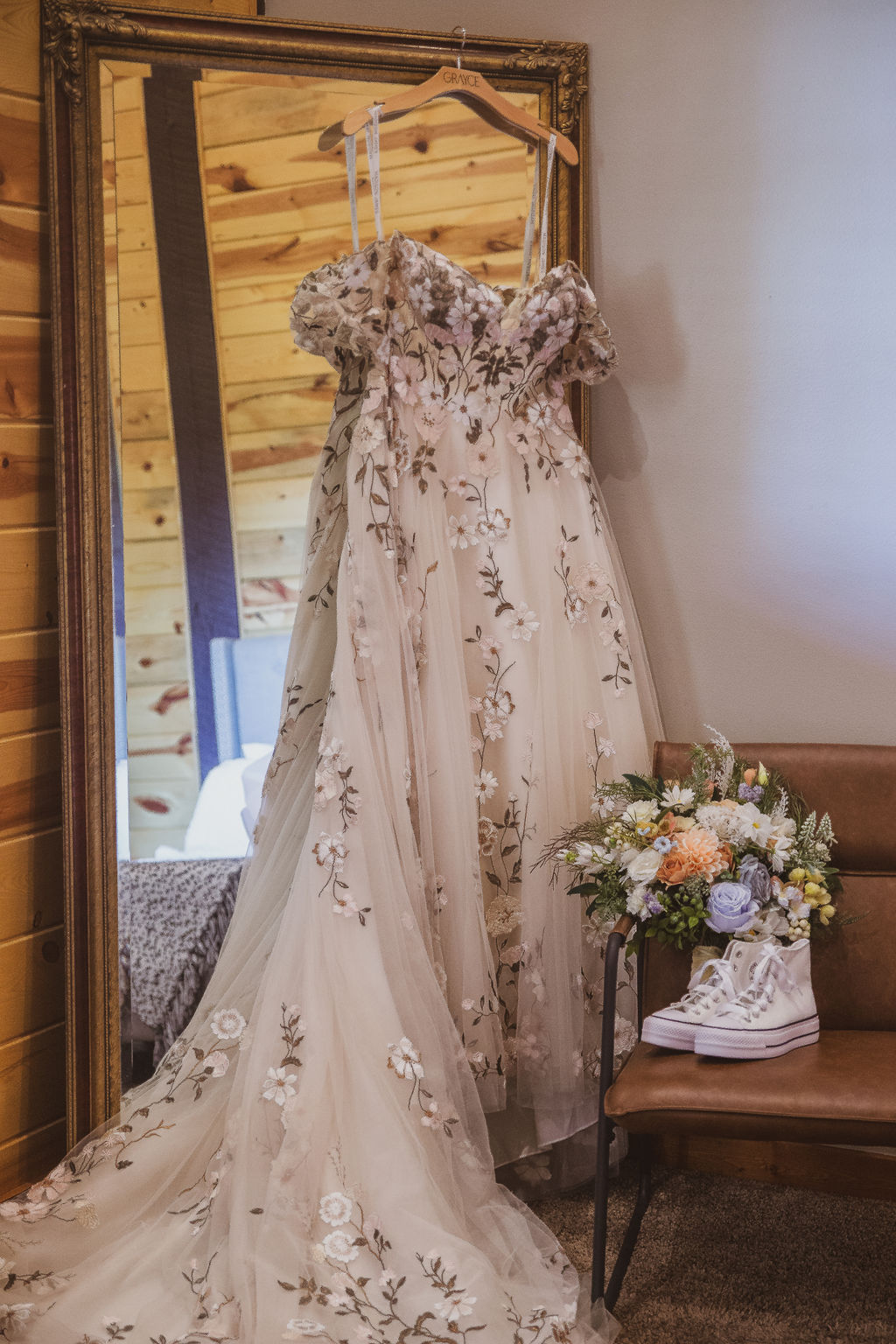 Jen & Alec's floral-embroidered gown hangs by a gold mirror with bouquet and sneakers at Covey Creek