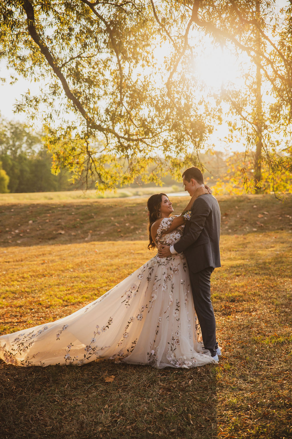Jen & Alec embrace under a tree at golden hour in her floral gown across a field at Covey Creek