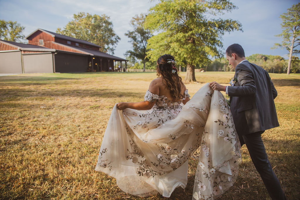 Jen & Alec walk toward the wooden barn as Alec carries her embroidered train at Covey Creek