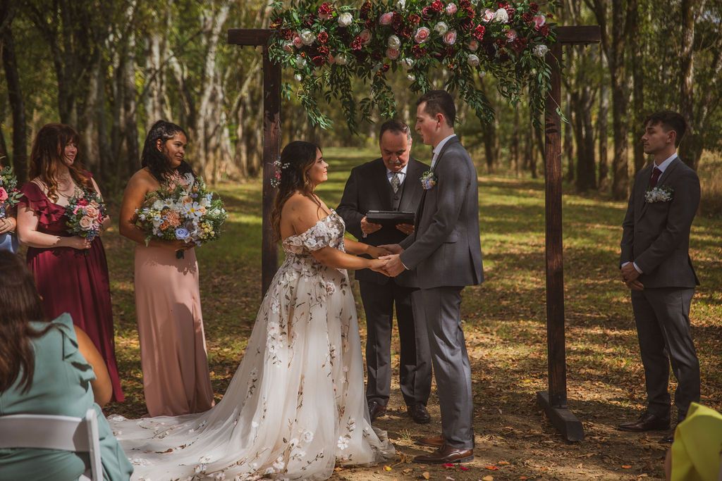 Jen & Alec hold hands exchanging vows under a floral arbor in the Tunnel of Love at Covey Creek
