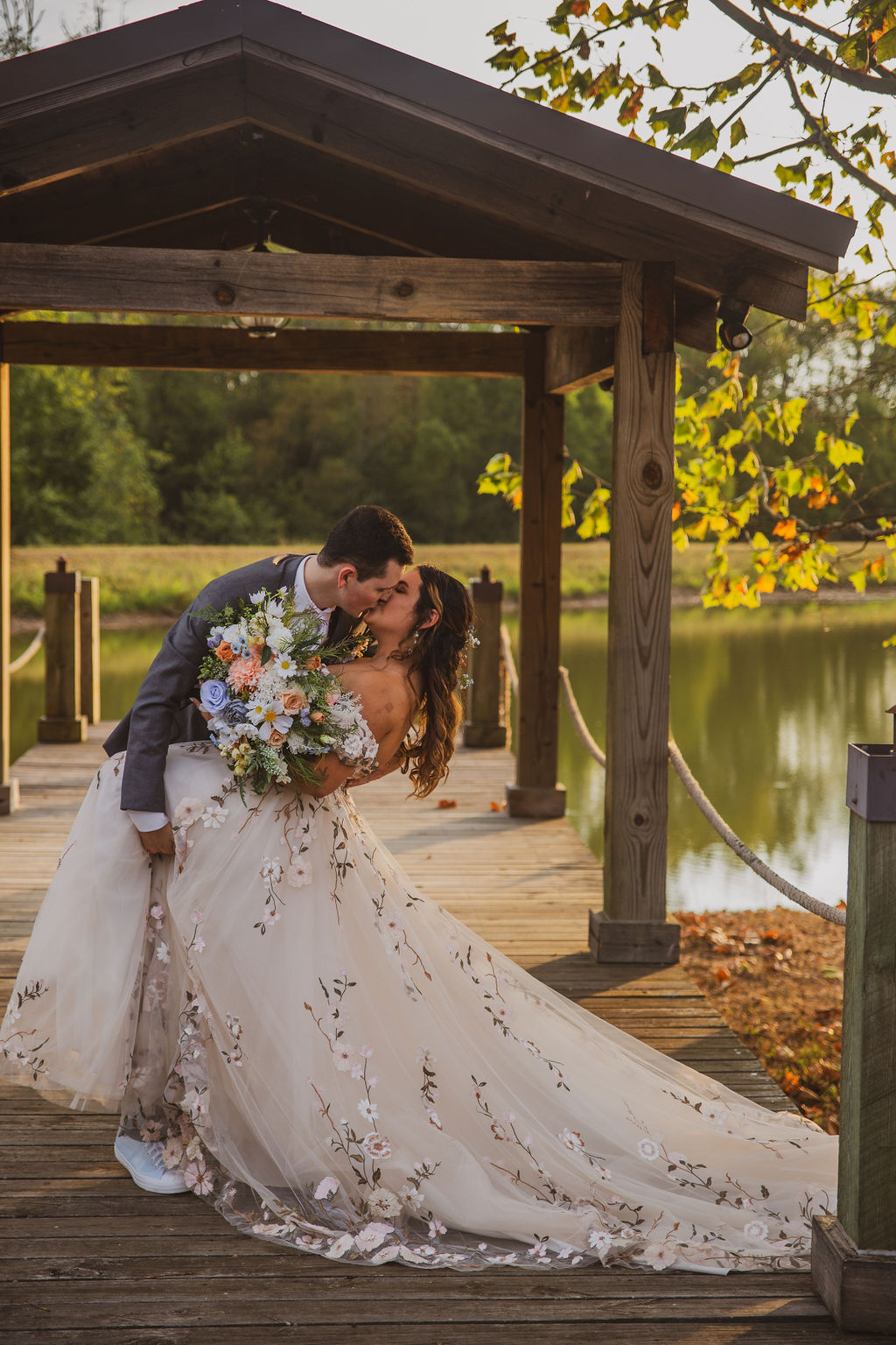 Jen & Alec share a dip kiss on the Pond dock pavilion, her floral train trailing at Covey Creek