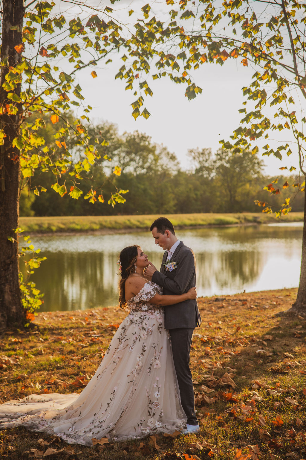 Jen & Alec gaze beside the Pond under autumn branches, her floral gown glowing at Covey Creek