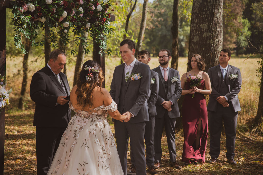 Jen & Alec stand under a rose arbor with wedding party in the Tunnel of Love at Covey Creek