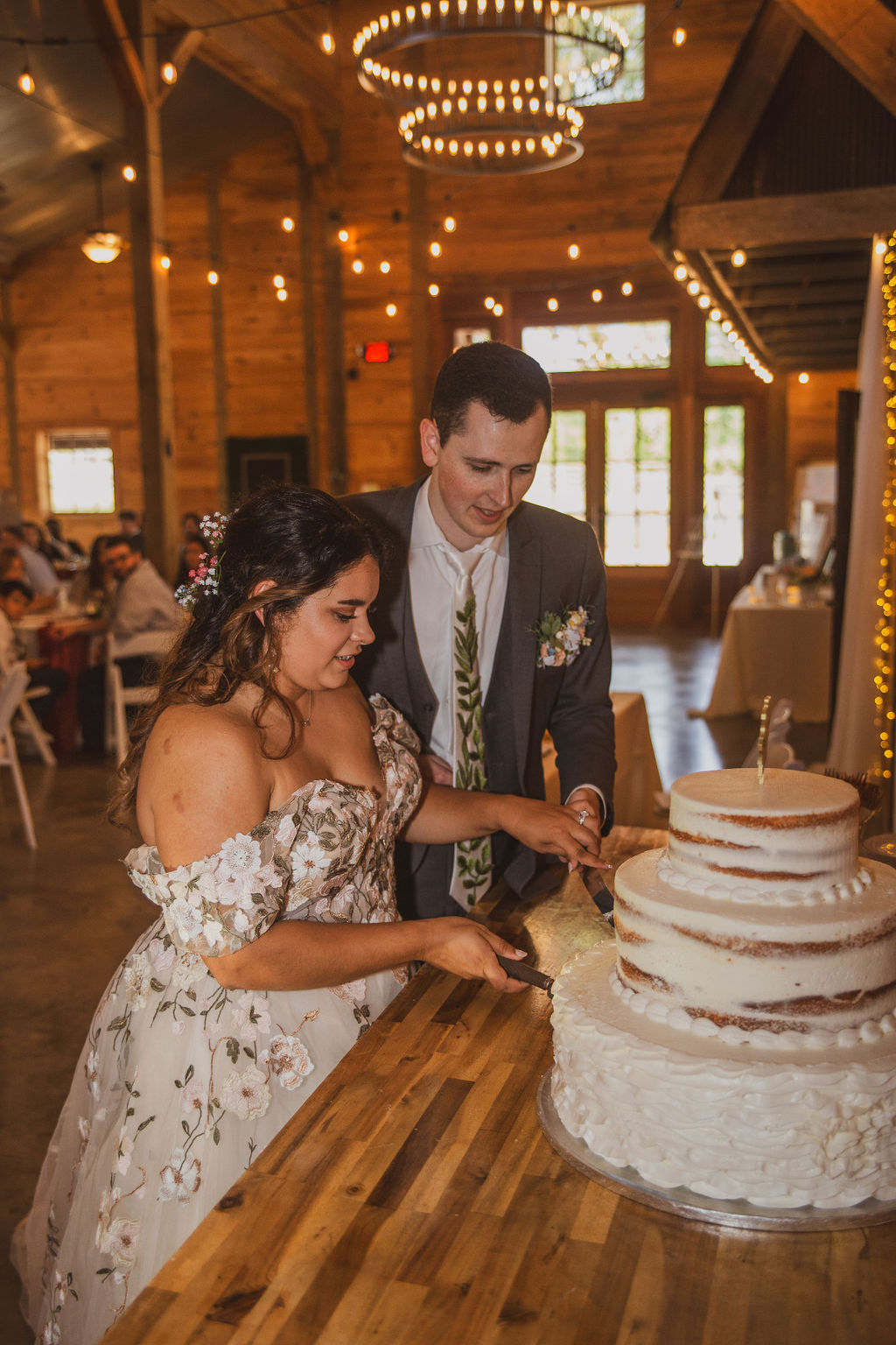 Jen & Alec cut their three-tier wedding cake under Edison chandeliers in the barn at Covey Creek