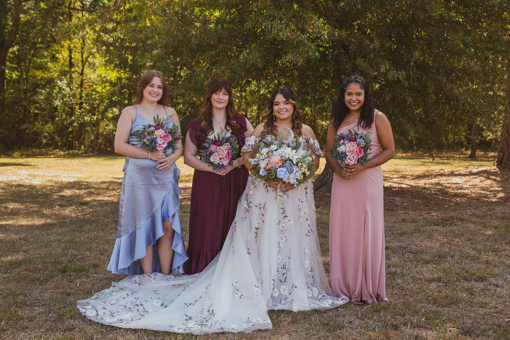 Jen & Alec's bridal party smiles in blue, burgundy, and blush gowns in a field at Covey Creek