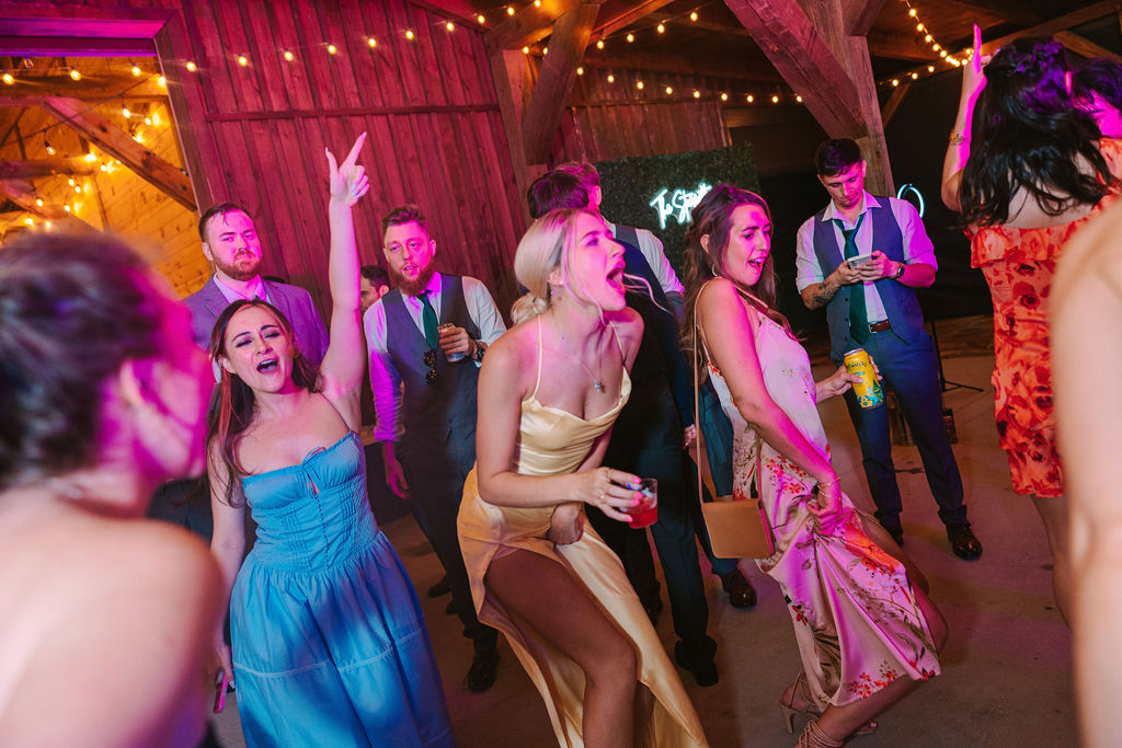 Guests dance at Kate & Josh’s Covey Creek reception, singing under pink barn lights and a greenery wall