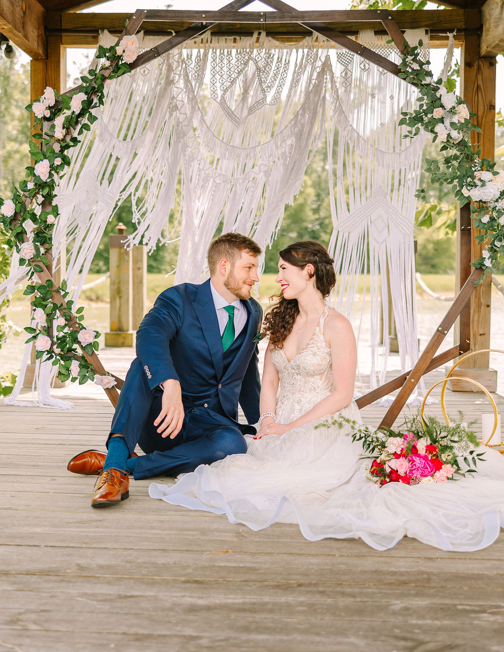 Kate & Josh sit cross-legged on Covey Creek’s Pond dock before a macrame hexagonal floral arch